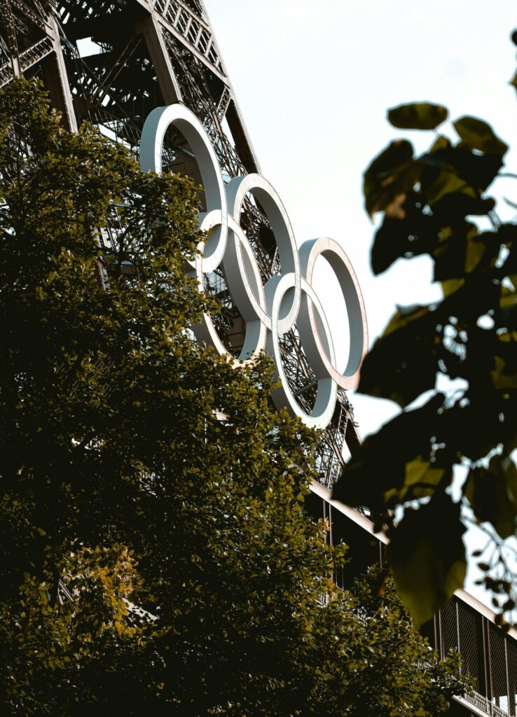 Close-up of the Eiffel Tower adorned with Olympic Rings, surrounded by lush greenery in daylight.